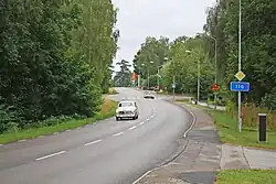Picture of a road surrounded by trees in Fridafors, Sweden. One car is driving towards the camera, while another is driving away through the intersection.