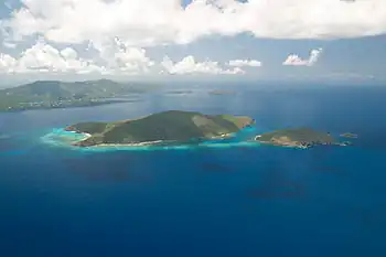 A small and a medium-sized island size covered in vegetation sit in the middle of a sea. The water is mostly dark blue, but lighter blue near the islands' shores. In the distance, to the left, a large island is visible.