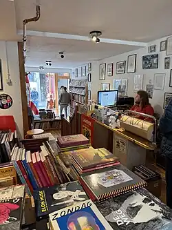 The Galerie Lambiek store interior showing a stack of comic books in the foreground with a man sitting behind a cash register in the background.