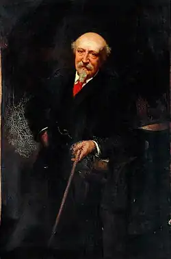 A colour painting of a mostly bald man leaning against a desk. He is holding a cane in his left hand and has his right hand on his hip. He is wearing a long coat and red jumper and looks stern and directly forward.