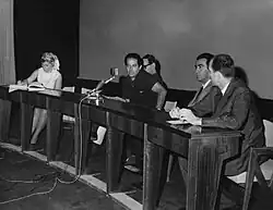 The picture is in black and white and it depicts four men and a woman behind a wide desk. In the center sits director Gillo Pontecorvo with a microphone in front of him.