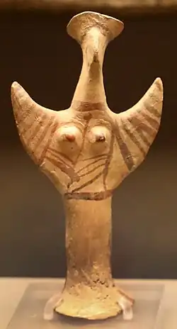 Small terracotta humanoid figurine: its arms are raised to the sky, as if praying. It has prominent breasts and wears a round hat known as a "polos".