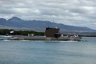 Side view of submarine with sailors on deck, at sea in front of a hilly coast