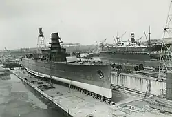 The unpainted cruiser in a shipyard, adjacent to the water and on a slipway. The entire ship is out of the water. While the ship's shape is present, she lacks all guns, sensors, funnel, and parts of the aft superstructure.