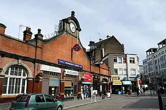 A brown-bricked building with a rectangular, blue sign reading "HAMMERSMITH STATION" in white letters all under a grey sky