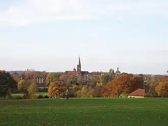 A view of Hampstead Heath across a grass covered field towards trees and a distant church tower.