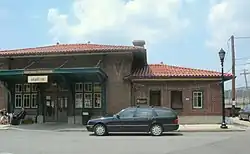 Entrance to a train station, signs read "Hastings-on-Hudson", "Station Cafe", "MTA Metro-North Railroad". Two people are sitting out front drinking coffee, and a black Mercedes wagon is parked.