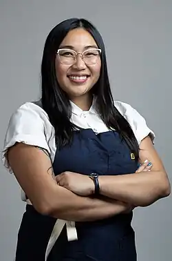 Nguyen is wearing an apron and smiling at the camera. She wears glasses and has long, straight hair.