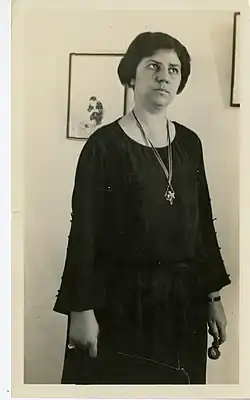 A white woman standing indoors, with short dark hair, wearing a loose-fitting dark dress with a pendant necklace