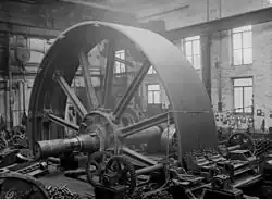 Flywheel for a large textile mill engine 1900, set up to machine grooves for the rope drives simultaneously. The saddle with two tool posts to the front. The wheel is rotated by two pinions driving via the cast-in barring gear teeth in the flywheel rim. Temporary wedges are securing the spokes to the hub of the wheel. A travelling crane behind and above.[80][120]