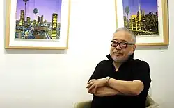 Hiroshi Nagai in 2020, standing in front of two of his colorful cityscape paintings.