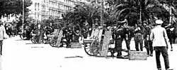 Black and white photograph of people standing with four wheeled guns. There are buildings and palms in the background.