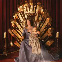 Halsey wears a blue-gray dress while sitting on a gold throne and breastfeeding a baby draped in beige cloth.