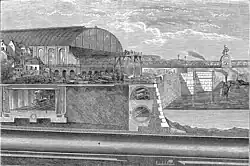 Illustrated section showing below ground structures of a riverside embankment including a covered railway line with steam train, riverside wall with sewer and pipes running behind. A large, glass roofed railway station sits in the left middle distance adjoining a railway bridge that crosses the river. Boats ply the water and tiny figures are engaged in construction work with a raised scaffold in the centre.
