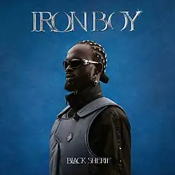 At the top is the title of the album, Iron Boy, in all caps with an iron-like outlining. The rest of the image shows Black Sherif in darkish clothing at the center to bottom of the image.