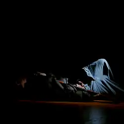 In darkness, a person lays on the ground with a gun laying across their chest, highlighted only by a faint spotlight