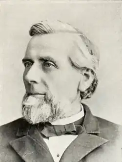 Head and shoulders photograph of a man in an overcoat. He has a salt and pepper goatee and has his hair combed to the side. He wears a calm expression and is looking slightly to the right.