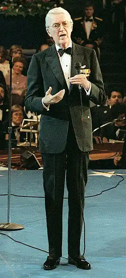 An elderly Stewart standing in a tuxedo on a stage, holding a microphone