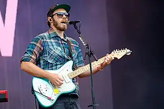 James Vincent McMorrow wearing a checkered shirt, dark blue pants, and dark sunglasses, playing electric guitar and singing into a microphone onstage