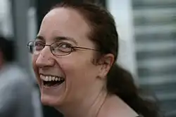 Photograph of Jeni Tennison. She is looking back over her shoulder toward the camera laughing. She is a white female with long dark hair, tied back, and is wearing glasses.