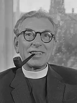 Black-and-white photo of John Collins wearing glasses, clerical collar, and a suit jacket while smoking a pipe