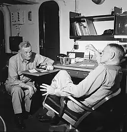 Black and white photograph of two men seated in an office. Both are wearing khakis