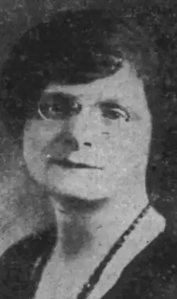 A white woman with dark hair, wearing pince-nez glasses and a strand of dark beads