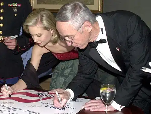 A young woman and an older man, both dressed in formal wear, leaning over to sign a poster.