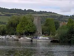 Looking across water to moored boats. Beyond them is a stone chimney surrounded by trees, with hills in the distance.