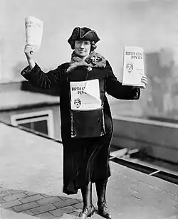 Awoman standing on a street, holding flyers for sale.