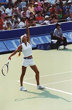 Anna Kournikova playing tennis in white outfit. Left hand is extended as if she has just tossed a ball and right hand is cocking back for the serve.