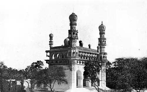 A black and white image of a small mosque amidst thick trees. The mosque is richly decorated with two large minarets in the front, and some smaller minarets and a dome only partially visible at the rear.