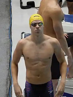 Léon Marchand shown from about the thigh up in swim trunks, no shirt, black swim goggles, and a yellow swim cap.