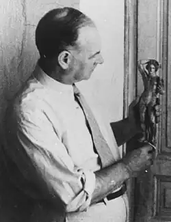 Shear, with a slightly receding hairline, holds and admires a classical statuette of Apollo. He is wearing a white shirt and a tie.