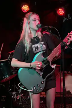 Full-body shot of Lizzie Esau, a young white, woman with bleached hair, wearing a dark T-shirt, shorts and fishnets, singing and playing an electric guitar.