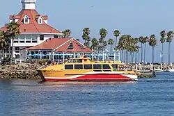 Long Beach Transit's AquaLink II water taxi approaches Alamitos Bay Landing. Palm trees and a restaurant are visible in the background.