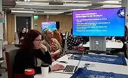 A computer screen reading "What can university departments do to help?" on a desk with people sitting at it