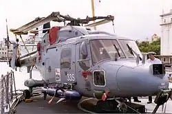 A light blue helicopter on a ship's flight deck with its rotor blades folded away. A large dark blue missile is attached to its right side and the number 335 is written in white on its door.