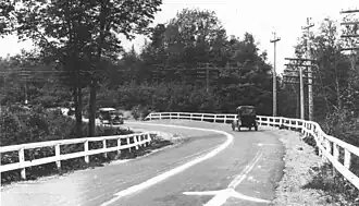 An old photograph of a bend in a road surrounded by trees and power poles. There are wooden guardrails on either side of the road, with a white-painted centre line separating the two lanes of traffic. Two old cars are approaching the curve which also has arrows to denote the direction of traffic.