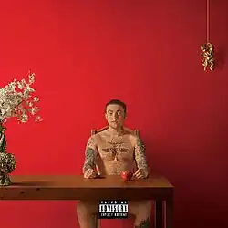 A red colored room. Mac sits at a table, completely naked, with his hands on the table and an apple next to his left hand. On the left side, there is a decorative vase with flowers inside, and on the top-right, a small golden figurine hangs from a string. A Parental Advisory label covers Mac's groin area.