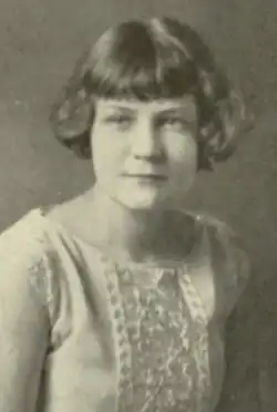 A young woman with bobbed blonde hair, wearing a sleeveless white dress with a lace detail in front