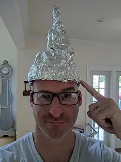 A man points to a large tin foil hat he is wearing while looking at the viewer with a smug expression.