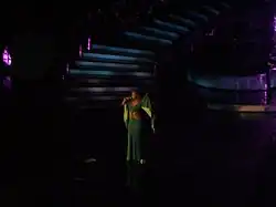 A woman is performing a song on stage. She is wearing a greenish dress.