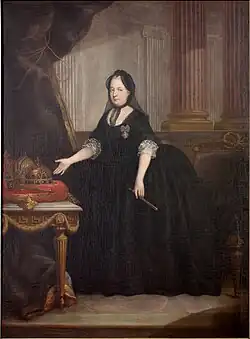 An elderly woman in a black dress and a black cap and weil, standing next to a table displaying the crowns and regalia of Hungary and Bohemia. With her right hand, she is pointing towards the crown, in the left, she holds a gilded fan.