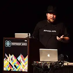 A bearded White man wearing a dark shirt and hat stands before a computer situated on a table next to a podium.