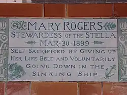 A tablet formed of two large tiles, bordered by green flowers in the style of the Arts and Crafts movement, and decorated with a stylised ship and anchor. The tablet reads "Mary Rogers, Stewardess of the Stella, March 30, 1899, Self sacrificed by giving up her life belt and voluntarily going down in the sinking ship".
