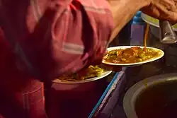 An image of a food vendor preparing masala puri. He has two plates in hand with the dry ingredients of the dish, upon which he pours the spiced gravy.