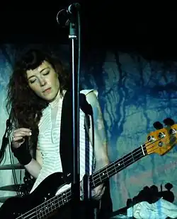 A female musician performing with a bass guitar against a black and blue backdrop. A microphone is visible in front of her.