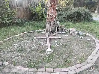 A folk art memorial at the base of a tree. A cross made of stones is laid out on the ground within a circle of bricks. The tree trunk behind it is adorned with small, handmade plaques and notes.