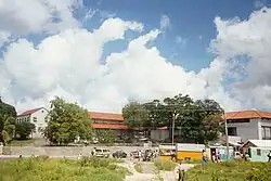 In the foreground there is a large building with red roofs. There are trees surrounding the it. The sky is blue with some white clouds.